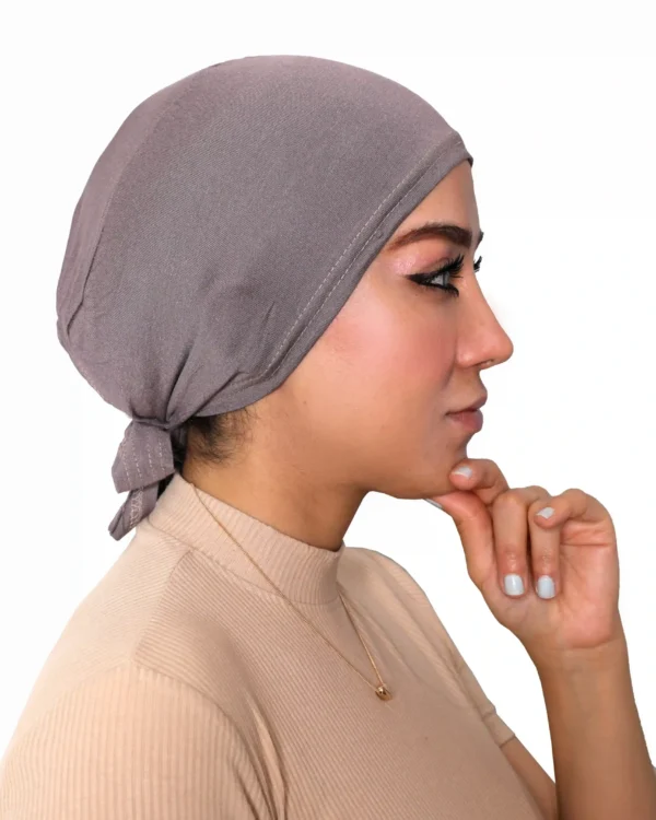 A woman models a Chic Grey Turban Cap for Women, viewed from the side and looking down, highlighting the gentle pleating and single line of stitching above the brow.