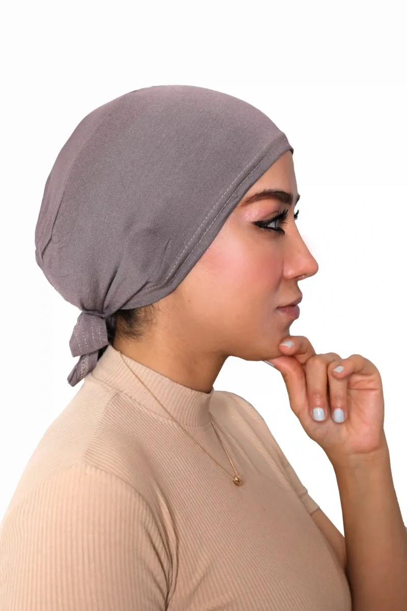 A woman models a Chic Grey Turban Cap for Women, viewed from the side and looking down, highlighting the gentle pleating and single line of stitching above the brow.